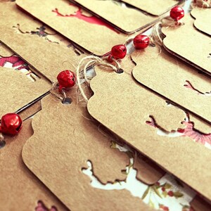 K&ouml;nnte beinhalten: Ein Stapel brauner Papier-Geschenk-Etiketten mit einer ausgeschnittenen Rentier-Silhouette. Die Etiketten sind mit Schnur und roten Gl&ouml;ckchen zusammengebunden.