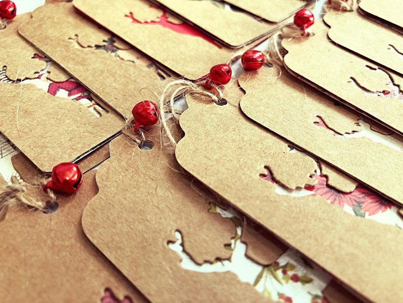 K&ouml;nnte beinhalten: Ein Stapel brauner Papier-Geschenk-Etiketten mit einer ausgeschnittenen Rentier-Silhouette. Die Etiketten sind mit Schnur und roten Gl&ouml;ckchen zusammengebunden.