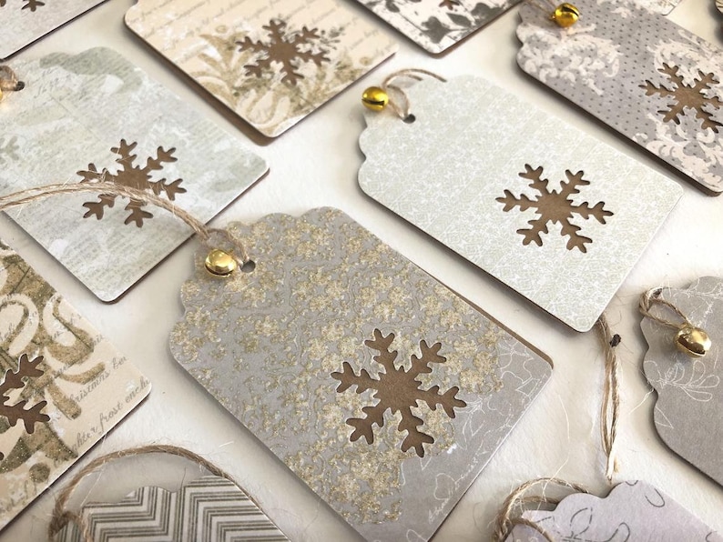 May include: A set of 10 gift tags with a snowflake design. The tags are made of brown cardstock and have a white background with a gold glitter finish. Each tag has a gold bell attached to a twine string.