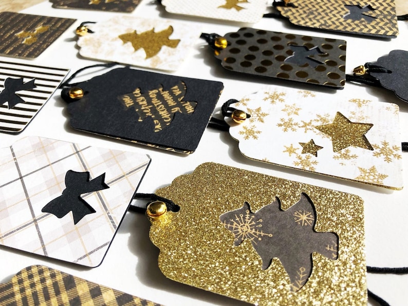 Pu&ograve; includere: Una collezione di etichette regalo di Natale nere e dorate con vari disegni, tra cui fiocchi di neve, stelle, fiocchi e alberi di Natale. Le etichette sono realizzate in carta e hanno una finitura glitter.