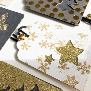 Pu&ograve; includere: Etichetta regalo bianca con fiocchi di neve e stelle dorate glitter. L'etichetta ha un laccio nero.
