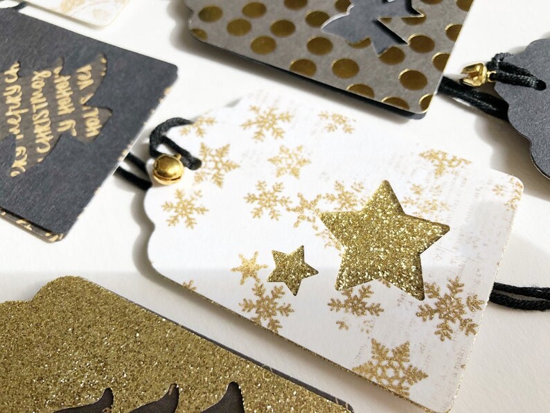 Pu&ograve; includere: Etichetta regalo bianca con fiocchi di neve e stelle dorate glitter. L'etichetta ha un laccio nero.