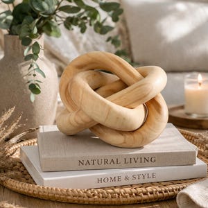 May include: A light-coloured wooden knot sculpture sits on two books with the titles "NATURAL LIVING" and "HOME & STYLE". A vase with greenery and a lit candle are in the background. The sculpture is a decorative accent piece.