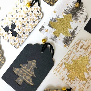 Pu&ograve; includere: Una collezione di etichette regalo di Natale con vari design. Le etichette sono realizzate in cartoncino bianco con accenti di glitter dorati e presentano diversi design di alberi di Natale. Alcune etichette hanno uno sfondo nero con un albero di glitter dorato ritagliato.