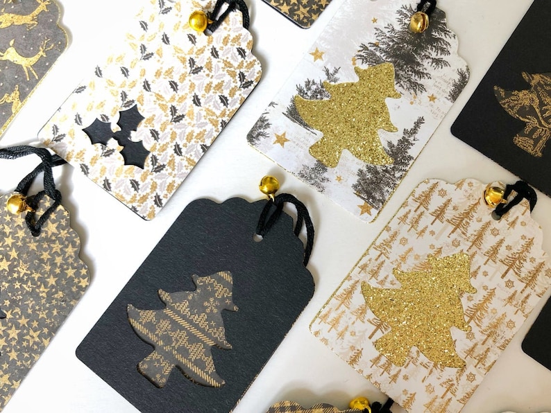 Pu&ograve; includere: Una collezione di etichette regalo di Natale con vari design. Le etichette sono realizzate in cartoncino bianco con accenti di glitter dorati e presentano diversi design di alberi di Natale. Alcune etichette hanno uno sfondo nero con un albero di glitter dorato ritagliato.