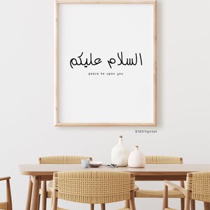 May include: A framed print with the Arabic phrase "Assalamu Alaykum" which translates to "Peace be upon you" in English. The print is hanging on a white wall above a wooden table with woven chairs.