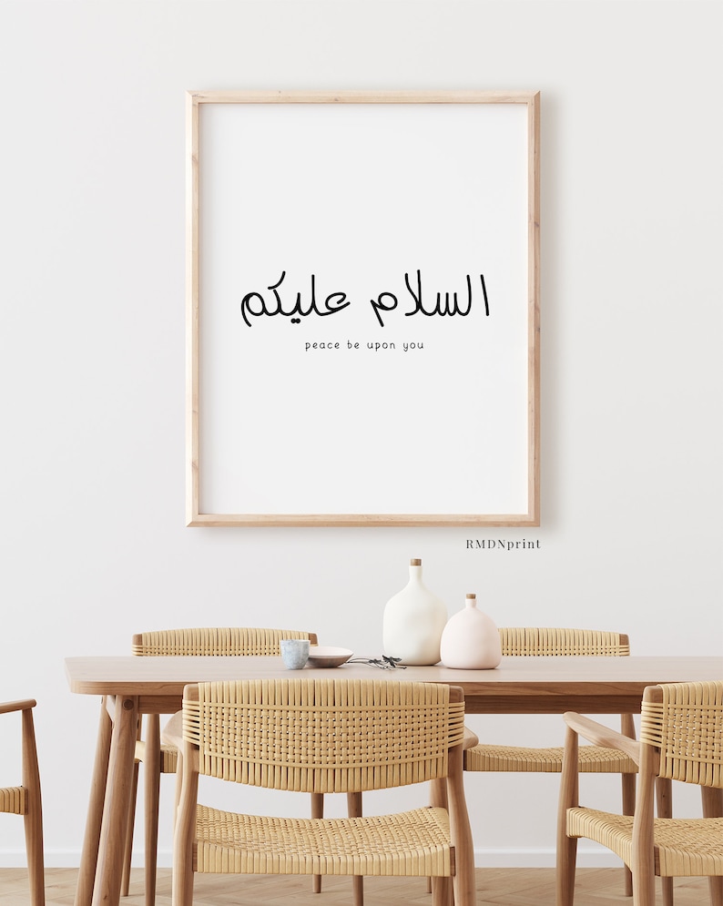May include: A framed print with the Arabic phrase "Assalamu Alaykum" which translates to "Peace be upon you" in English. The print is hanging on a white wall above a wooden table with woven chairs.