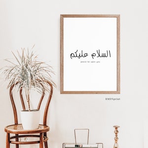 May include: A framed print with the Arabic phrase "Assalamu Alaykum" which translates to "Peace be upon you". The print is hanging on a white wall with a potted plant and a wooden chair in the foreground.
