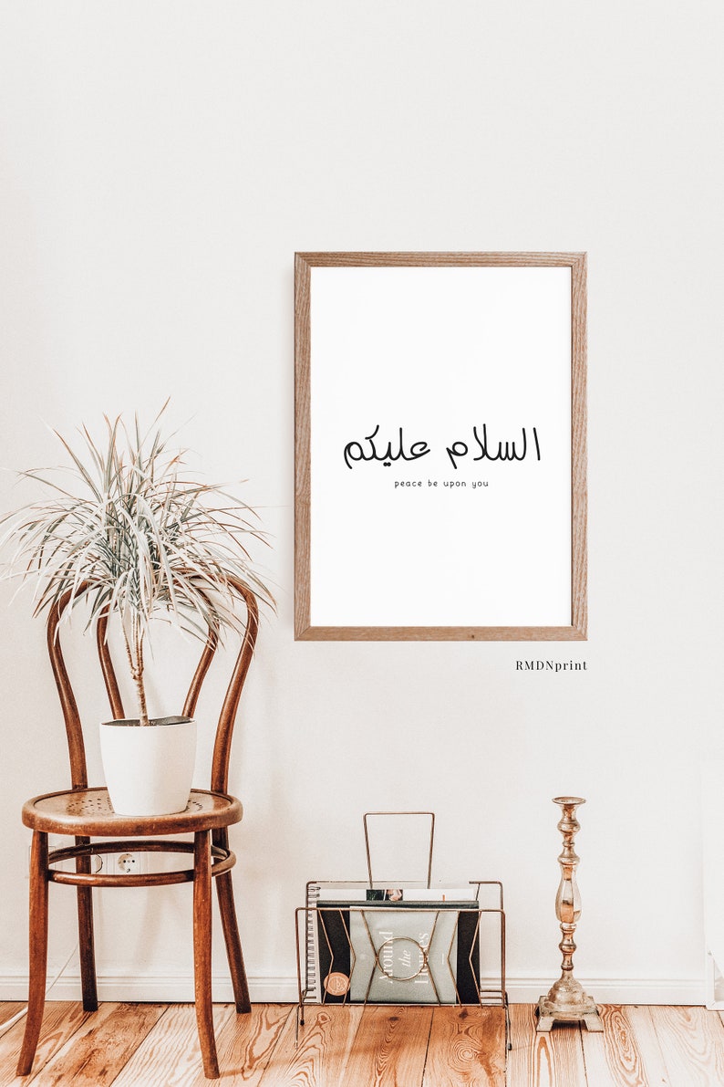 May include: A framed print with the Arabic phrase "Assalamu Alaykum" which translates to "Peace be upon you". The print is hanging on a white wall with a potted plant and a wooden chair in the foreground.