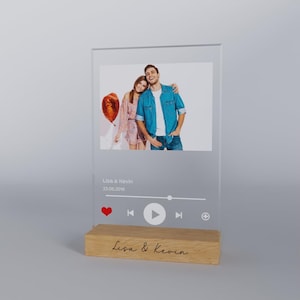 Peut inclure: Un cadre photo en acrylique transparent avec une base en bois. Le cadre présente une photo d'un couple avec un ballon rouge, un bouton de lecture et le texte "Lisa & Kevin 23.06.2019". La base porte le texte "Lisa & Kevin" écrit en cursive.