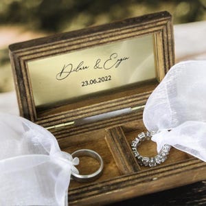 May include: A wooden ring box with a clear lid and a gold interior. The lid is engraved with the text "Dilan & Eryn 23.06.2022". Two rings are inside the box, one is a simple band and the other is a diamond ring.