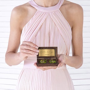 May include: A wooden ring box with a gold inscription and two gold rings inside. The box is open and the rings are visible. The box is being held by a person wearing a pink dress.