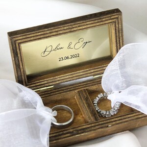 May include: A wooden ring box with a gold interior and a white satin ribbon. The box is engraved with the names "Dilara & Ergin" and the date "23.06.2022".