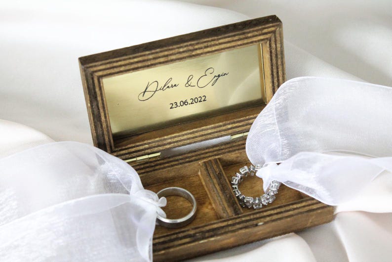 May include: A wooden ring box with a gold interior and a white satin ribbon. The box is engraved with the names "Dilara & Ergin" and the date "23.06.2022".