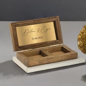 May include: A wooden ring box with a gold-colored plaque on the lid. The plaque has the names "Dilara & Ergin" and the date "23.06.2022" engraved on it.