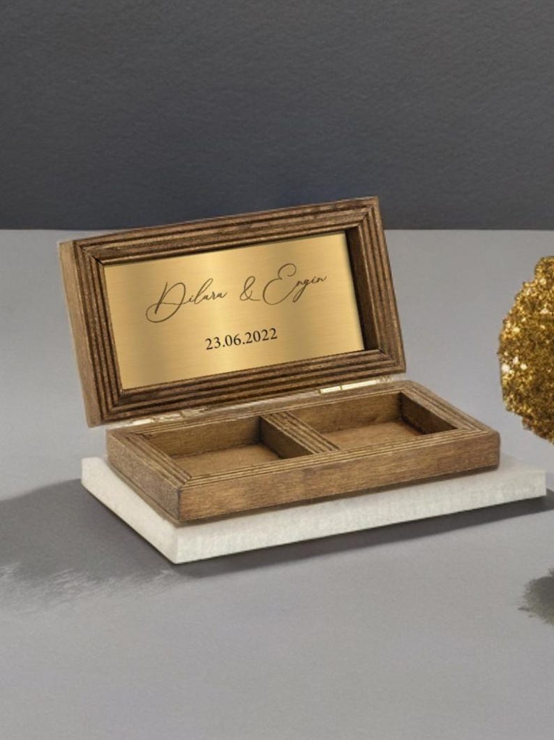 May include: A wooden ring box with a gold-colored plaque on the lid. The plaque has the names "Dilara & Ergin" and the date "23.06.2022" engraved on it.