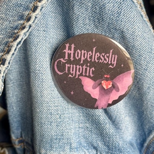 May include: A circular pinback button featuring a dark purple background with the text Hopelessly Cryptic in a stylized font. The design includes a moth-like creature with pink wings holding a small red heart, accented by tiny white stars.