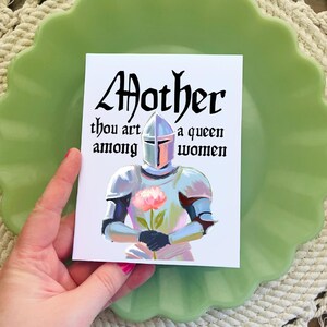May include: A white card with black text that reads "Mother thou art a queen among women". The card features a painting of a knight in silver armor holding a pink flower.