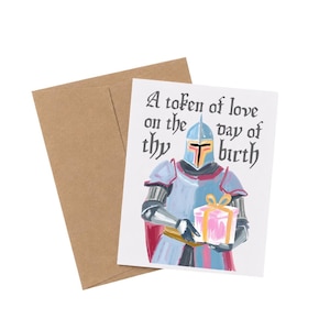 May include: A white greeting card with a brown envelope featuring a painted illustration of a knight in blue and silver armor holding a pink gift box with a gold ribbon. The text reads A token of love on the day of thy birth in black font.
