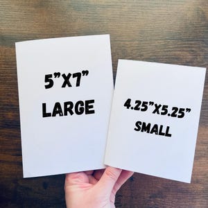 May include: Two white rectangular cards with black text. The larger card reads "5"X7" LARGE" and the smaller card reads "4.25"X5.25" SMALL". The cards are held against a brown wooden background.