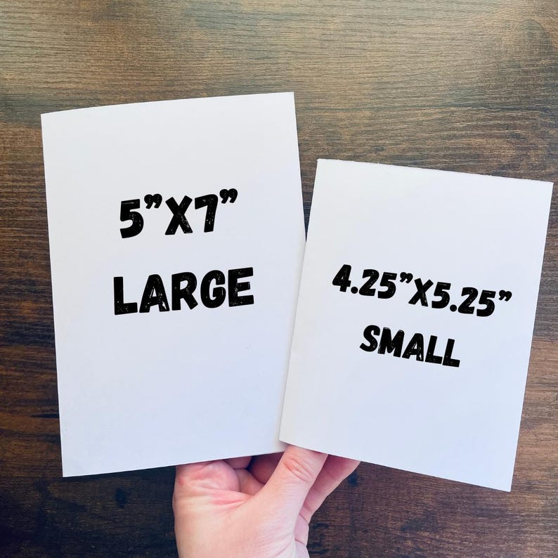 May include: Two white rectangular cards with black text. The larger card reads "5"X7" LARGE" and the smaller card reads "4.25"X5.25" SMALL". The cards are held against a brown wooden background.