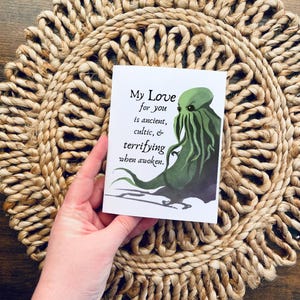 May include: A white greeting card with the text "My Love for you is ancient, cultic, & terrifying when awoken." features a green monster illustration. The card is held over a woven, circular placemat.