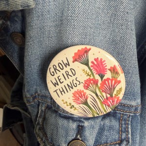 May include: A round pinback button with a cream-colored background featuring a botanical illustration of red and pink flowers with green stems. The text "GROW WEIRD THINGS." is printed in black capital letters on the left side of the button.