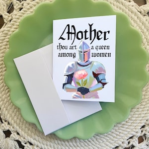 May include: A white card with a knight in silver armor holding a pink flower. The card has the text "Mother thou art a queen among women" in black.