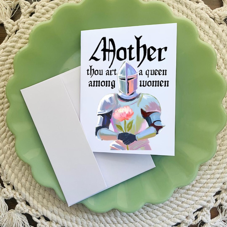 May include: A white card with a knight in silver armor holding a pink flower. The card has the text "Mother thou art a queen among women" in black.