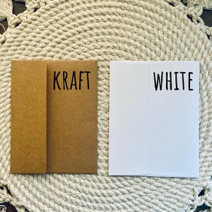 May include: Two folded paper cards, one kraft brown and one white, are displayed side-by-side. The kraft card has the word "KRAFT" printed in black, while the white card has "WHITE" printed in black. The cards are on a textured, off-white surface.