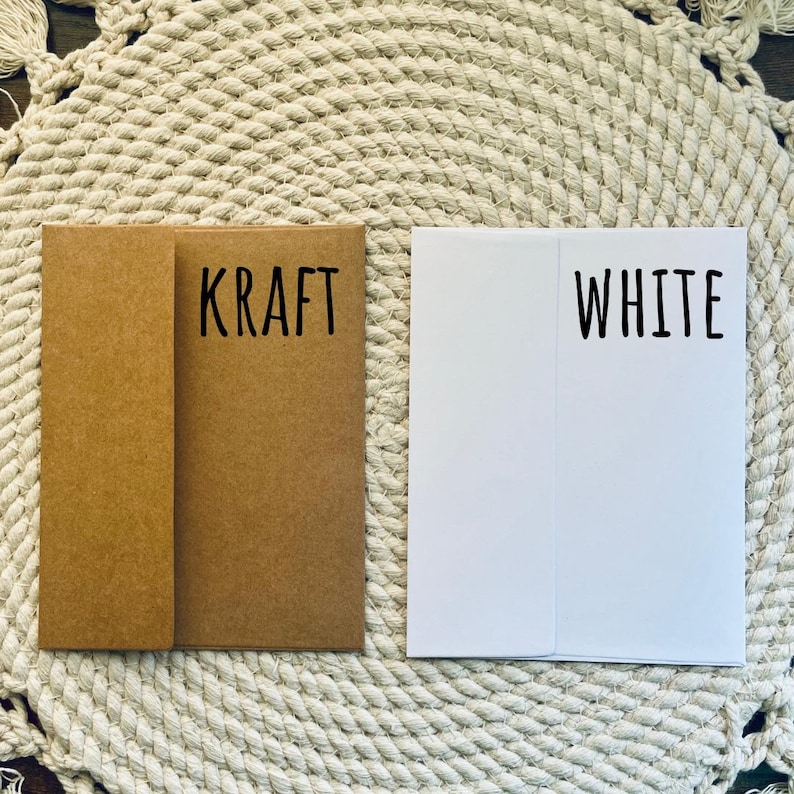 May include: Two folded paper cards, one kraft brown and one white, are displayed side-by-side. The kraft card has the word "KRAFT" printed in black, while the white card has "WHITE" printed in black. The cards are on a textured, off-white surface.