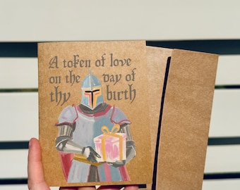 May include: A brown kraft paper greeting card featuring a painted illustration of a knight in silver armor holding a pink gift box with a yellow ribbon. The text reads A token of love on the day of thy birth in a dark, stylized gothic font.