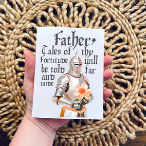 May include: A white card with a knight in silver armour holding a pink flower. The card has the text "Father, Tales of thy fortitude will be told far and wide."