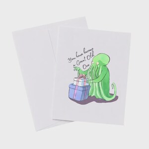 May include: A white greeting card and envelope featuring a green tentacled creature holding two stacked gift boxes with pink ribbons. The text on the card reads, You have become a Great Old One, written in a black cursive script above the illustration.