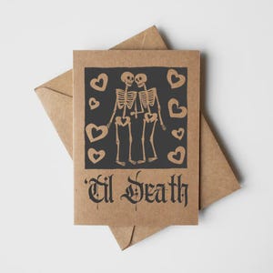 May include: A brown kraft paper greeting card featuring a black rectangular block print of two skeletons holding hands surrounded by heart shapes. Below the illustration, the text 'Til Death' is printed in a bold, black gothic-style font.