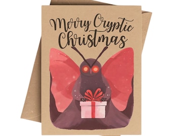 May include: A brown Christmas card with the text "Merry Cryptic Christmas" in black script. The card features a moth-like creature with red wings and glowing eyes, holding a gift box with a red bow. The card is inside a brown envelope.