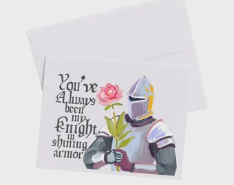 May include: A white greeting card and envelope featuring a painted illustration of a knight in silver armor holding a pink rose. The text on the card reads, You've Always been my Knight in shining armor, written in a black, stylized gothic font.