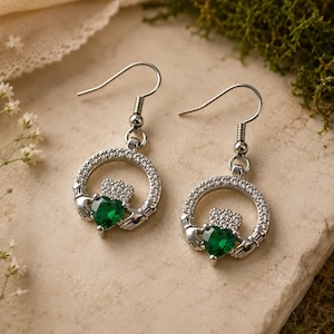 Può includere: Un paio di orecchini pendenti color argento con design Claddagh. Ogni orecchino presenta una pietra verde centrale a forma di cuore, una piccola corona sopra e due mani che sostengono il cuore, il tutto circondato da un cerchio di pietre chiare.