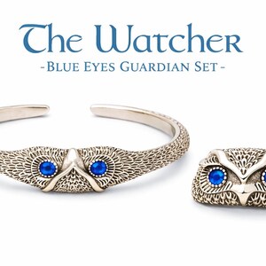 May include: Silver bracelet and ring set with an owl motif and blue gemstone eyes. The bracelet is open-ended, and the ring is an owl face. The text "The Watcher - Blue Eyes Guardian Set" is displayed above.