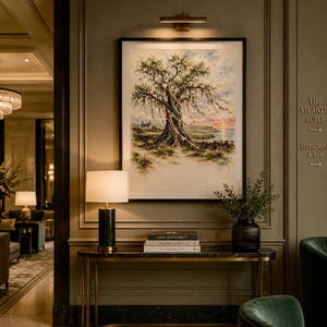 May include: A framed art print depicting a large tree with a landscape backdrop. The artwork hangs above a table with books and a lamp. The scene is set in a hotel or similar interior.