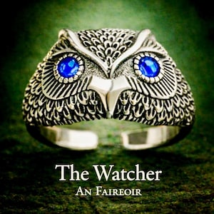 May include: A silver owl ring with blue gemstone eyes. The ring has detailed feather patterns and the text "The Watcher" and "An Faireoir". A distinctive piece of jewellery.