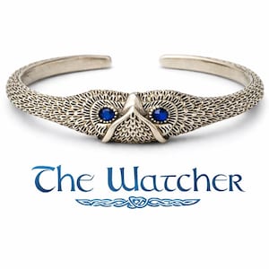 May include: Silver-toned cuff bracelet featuring an owl design with blue gemstone eyes. The bracelet has a textured, woven pattern and the text "The Watcher" in blue script.