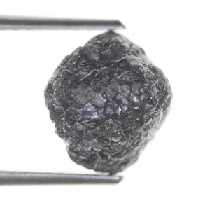 Pode incluir: Um diamante bruto cinza escuro, preso por uma pin&ccedil;a contra um fundo branco. A gema apresenta uma superf&iacute;cie irregular e texturizada, destacando sua forma natural e apar&ecirc;ncia n&atilde;o lapidada.