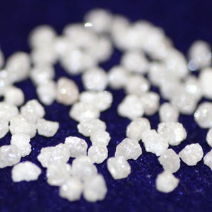 Op de afbeelding: Een close-up van een verzameling kleine, witte, ruwe diamanten die verspreid liggen op een donkerblauw oppervlak.