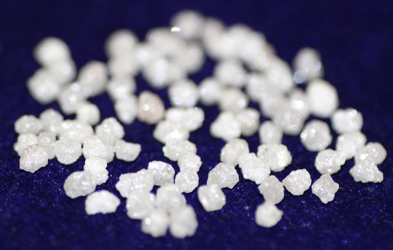 Op de afbeelding: Een close-up van een verzameling kleine, witte, ruwe diamanten die verspreid liggen op een donkerblauw oppervlak.