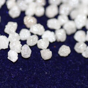 Op de afbeelding: Een close-up van een verzameling kleine, witte, ruwe diamanten op een donkerblauwe achtergrond.