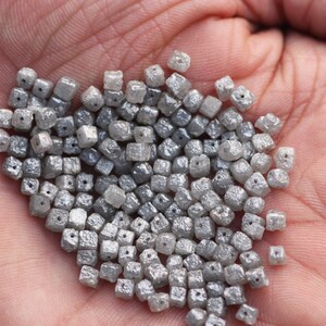 May include: A pile of small, gray, square-shaped diamonds with holes drilled through the center. The diamonds are loose and scattered on a hand.