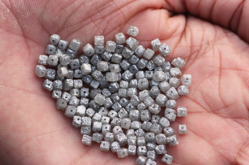 May include: A pile of small, gray, square-shaped diamonds with holes drilled through the center. The diamonds are loose and scattered on a hand.