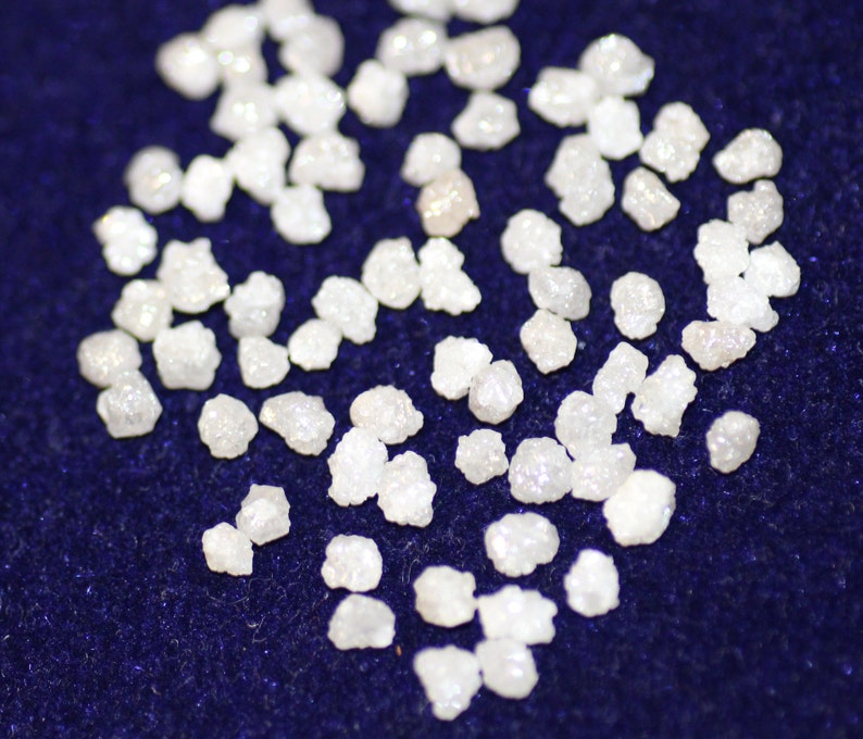 Op de afbeelding: Een close-up van een verzameling kleine, witte, ruwe diamanten die verspreid liggen op een donkerblauw oppervlak.