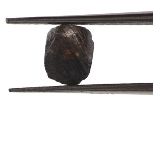 Puede incluir: Una piedra preciosa facetada de color gris oscuro sostenida por unas pinzas. La piedra preciosa es rugosa y tiene un aspecto natural sin pulir.